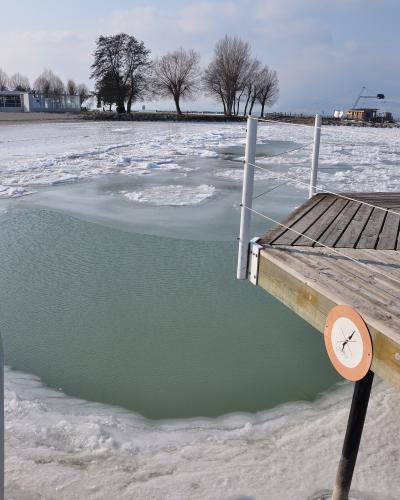 A frozen lake scene showing a wooden pier with white posts and a circular orange sign, with ice floes on the water and bare trees on the shore in the background.
