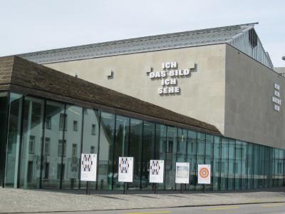 The image shows the exterior of a modern art museum building with a glass facade and a concrete upper section, featuring large German text on the wall reading "ICH DAS BILD ICH SEHE" and several exhibition posters in front.