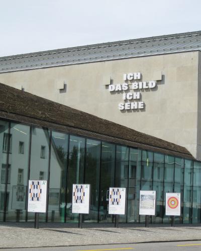 The image shows the exterior of a modern art museum building with a glass facade and a concrete upper section, featuring large German text on the wall reading "ICH DAS BILD ICH SEHE" and several exhibition posters in front.