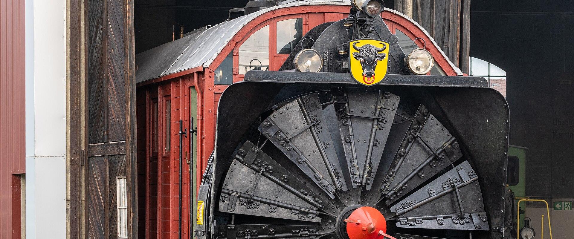 A large, black rotary snowplow locomotive with a distinctive yellow bull emblem on the front, emerging from a historic railway roundhouse or shed.