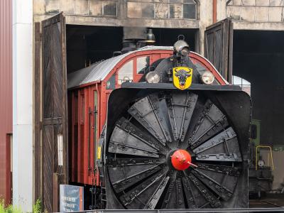 A large, black rotary snowplow locomotive with a distinctive yellow bull emblem on the front, emerging from a historic railway roundhouse or shed.