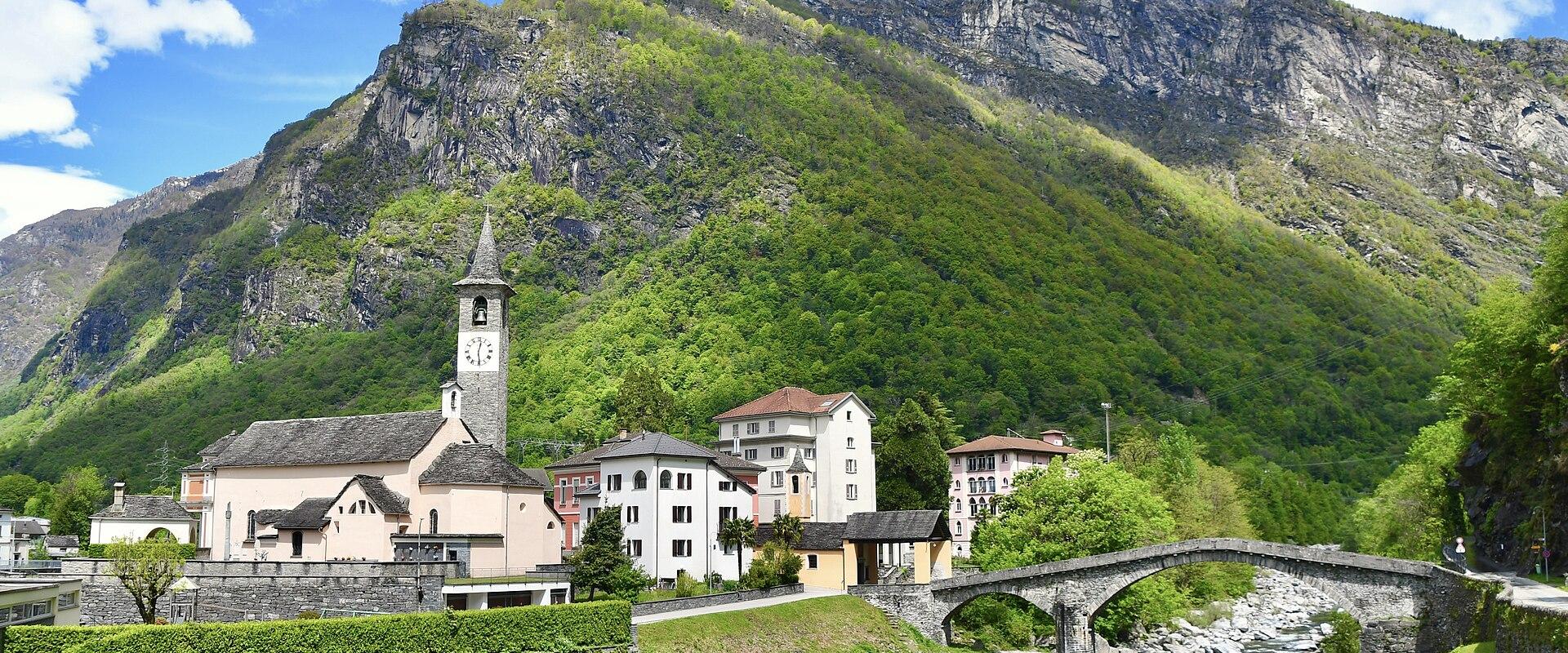 A scenic view of a Swiss village featuring a church with a clock tower, stone houses, and an arched stone bridge crossing a river at the foot of a steep, forested mountain.