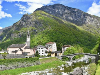 A scenic view of a Swiss village featuring a church with a clock tower, stone houses, and an arched stone bridge crossing a river at the foot of a steep, forested mountain.