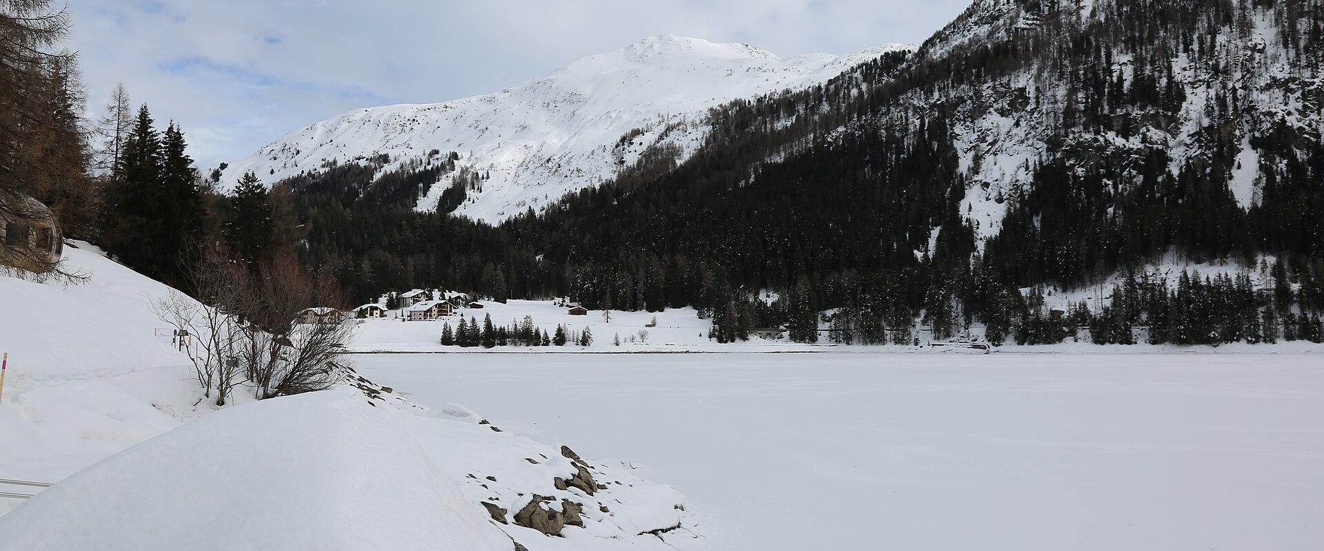 A wide, snow-covered frozen lake in a valley surrounded by steep, forested mountains and a few scattered chalets.