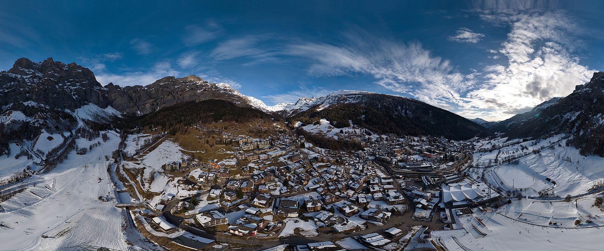 A 360-degree panoramic photograph of a snow-covered alpine village nestled in a valley surrounded by mountains, featuring numerous chalets and ski slopes.