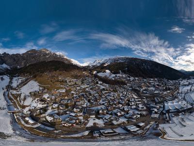 A 360-degree panoramic photograph of a snow-covered alpine village nestled in a valley surrounded by mountains, featuring numerous chalets and ski slopes.