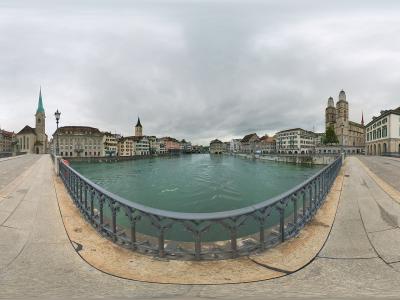 A 360-degree panoramic view from the center of a stone bridge spanning a wide river, featuring the Fraumünster church with its distinctive green spire on the left bank and the Grossmünster cathedral with its twin towers on the right bank.