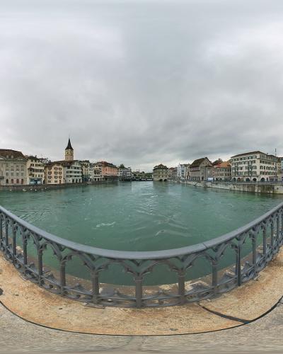 A 360-degree panoramic view from the center of a stone bridge spanning a wide river, featuring the Fraumünster church with its distinctive green spire on the left bank and the Grossmünster cathedral with its twin towers on the right bank.