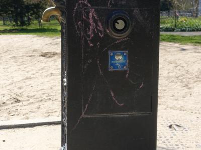 A close-up of a black metal public drinking fountain (borne-fontaine) with a brass spout, a coin slot, and a blue "ecopompe" sticker, set against a sandy ground and trees in the background.