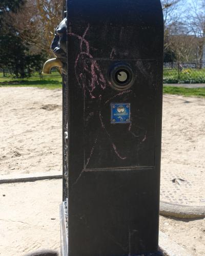 A close-up of a black metal public drinking fountain (borne-fontaine) with a brass spout, a coin slot, and a blue "ecopompe" sticker, set against a sandy ground and trees in the background.