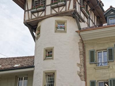 A low-angle shot of a distinctive white cylindrical tower with a projecting upper story featuring timber framing and flower boxes, situated between other buildings in an urban setting.