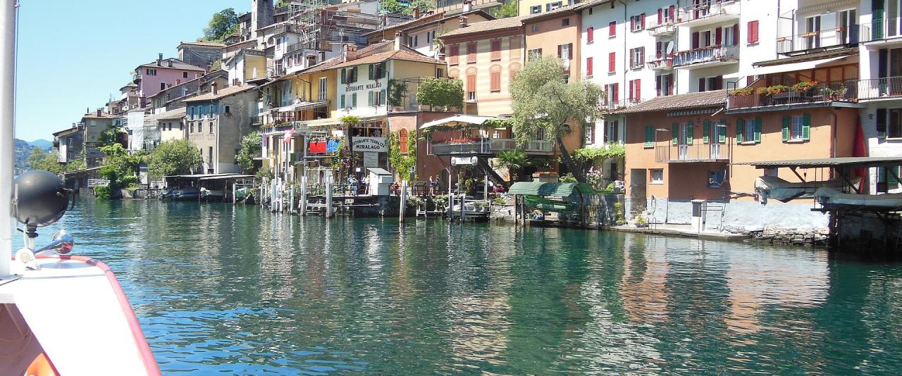 A view from a boat on a lake looking towards a hillside village with colorful buildings, small harbors, and a church tower.