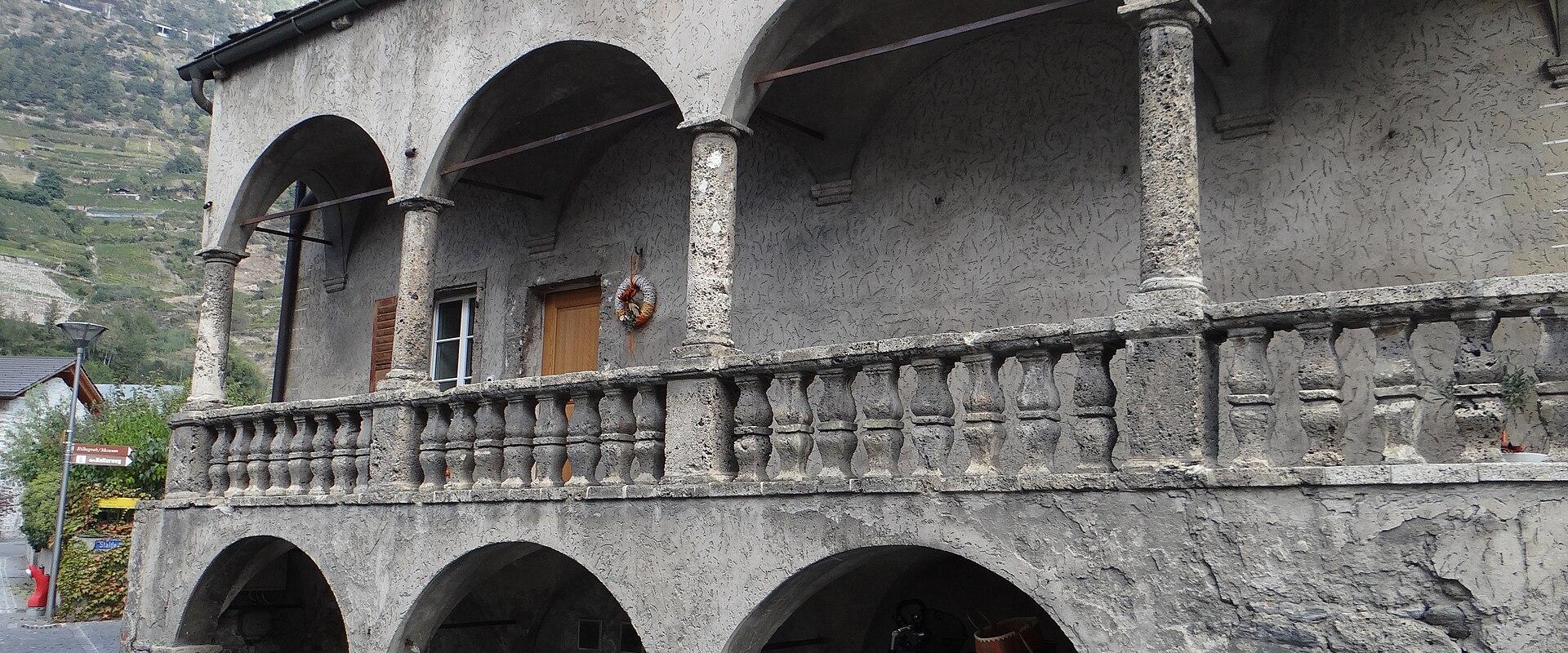 A two-story stone building with a covered arcade on the ground floor and an arched loggia on the upper floor, featuring a stone balustrade, set against a mountainous backdrop with a bicycle parked underneath.