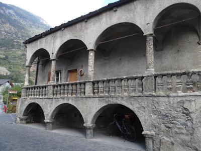 A two-story stone building with a covered arcade on the ground floor and an arched loggia on the upper floor, featuring a stone balustrade, set against a mountainous backdrop with a bicycle parked underneath.