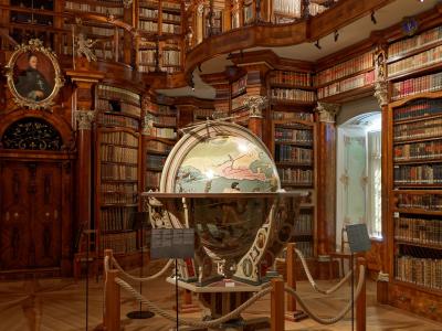 The image shows the interior of a grand, two-story baroque library hall with extensive wooden bookshelves, a painted ceiling, and a large terrestrial globe on display in the center.