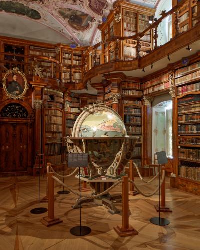 The image shows the interior of a grand, two-story baroque library hall with extensive wooden bookshelves, a painted ceiling, and a large terrestrial globe on display in the center.