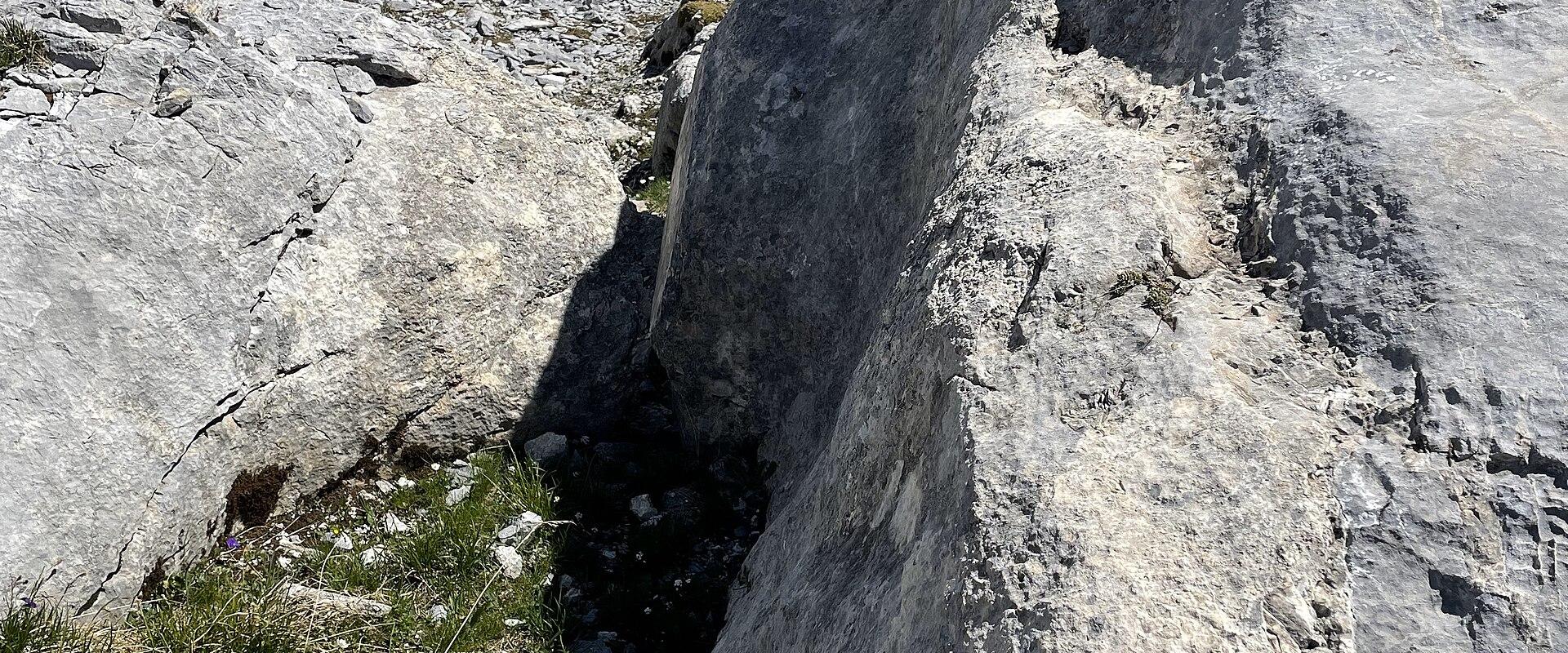 A close-up view of a large rock formation with a prominent vertical fissure or fault line, set against a backdrop of alpine slopes and distant snow-capped mountains.