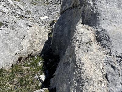 A close-up view of a large rock formation with a prominent vertical fissure or fault line, set against a backdrop of alpine slopes and distant snow-capped mountains.