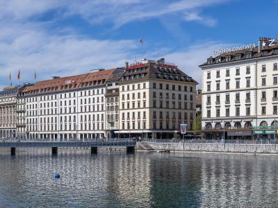 A view of the Quai des Bergues in Geneva, Switzerland, showing the Pont de la Machine bridge over the Rhône river with buildings featuring "VACHERON CONSTANTIN" and "ZENITH" signage on the right.
