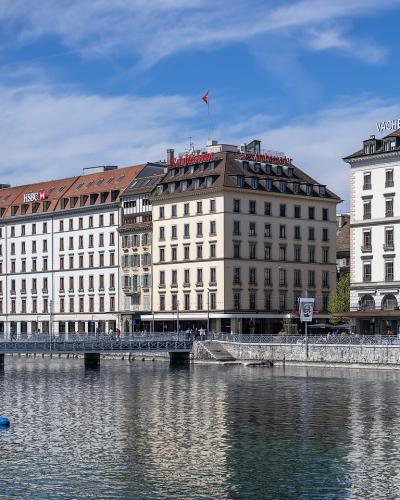 A view of the Quai des Bergues in Geneva, Switzerland, showing the Pont de la Machine bridge over the Rhône river with buildings featuring "VACHERON CONSTANTIN" and "ZENITH" signage on the right.