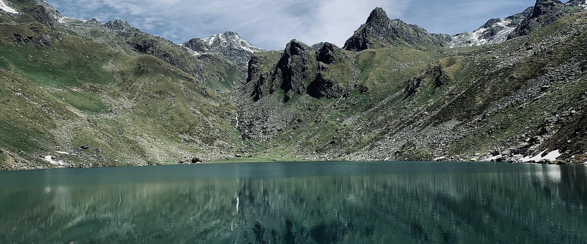 A high-altitude alpine lake surrounded by steep, rocky mountain slopes with patches of snow and green vegetation, reflecting the peaks in the calm water.