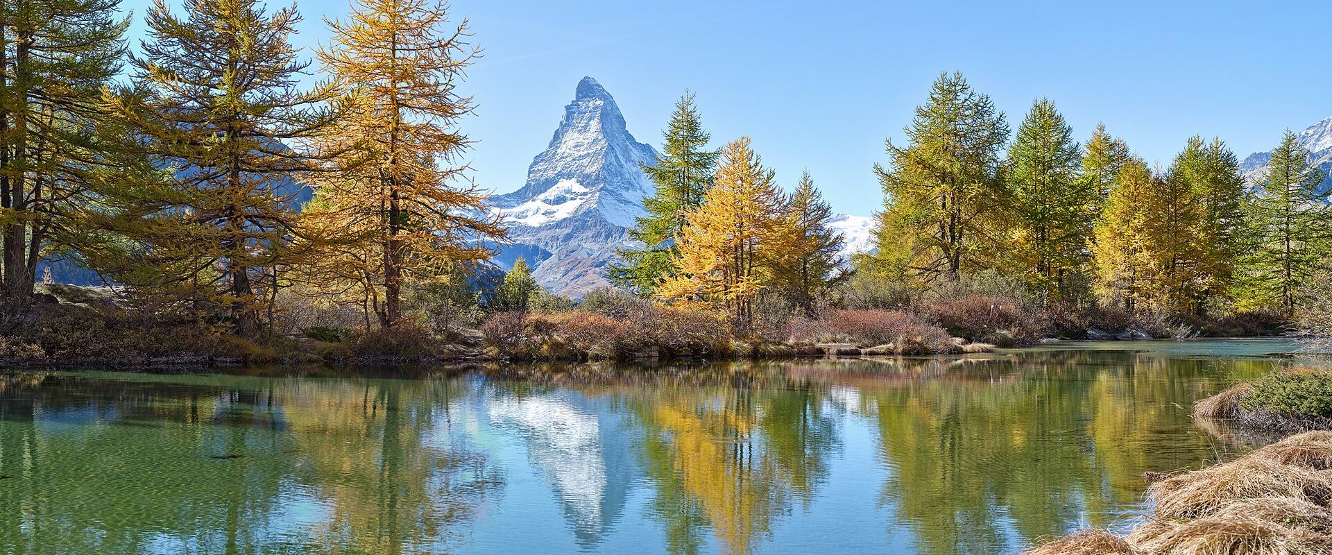 A landscape photograph showing the Matterhorn mountain reflected in a calm lake, framed by trees with yellow and green foliage under a blue sky.