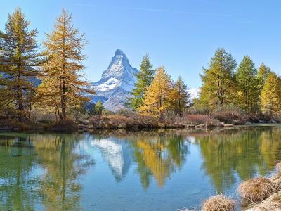 A landscape photograph showing the Matterhorn mountain reflected in a calm lake, framed by trees with yellow and green foliage under a blue sky.