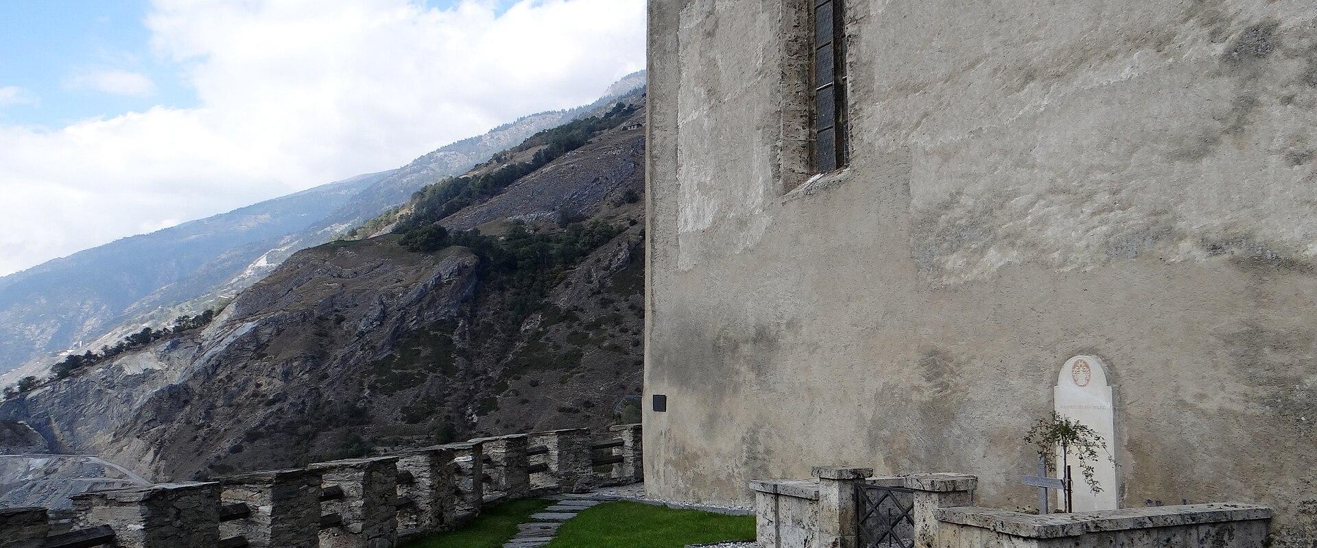 An exterior view of a weathered stone church wall with a tall arched window, a stone path, and a small memorial plot with a white headstone and cross in the foreground, set against a mountainous backdrop.