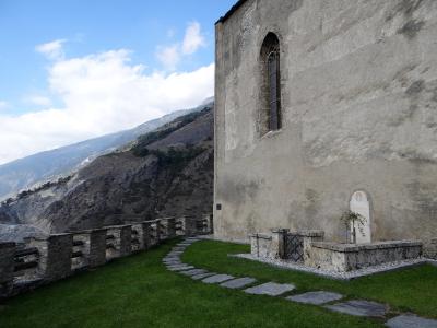 An exterior view of a weathered stone church wall with a tall arched window, a stone path, and a small memorial plot with a white headstone and cross in the foreground, set against a mountainous backdrop.