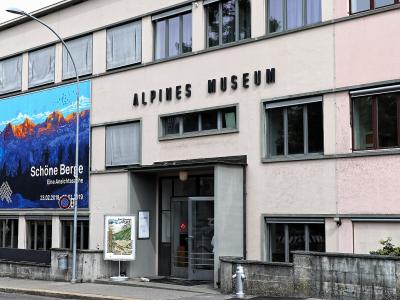 The image shows the exterior of a modern, multi-story building with a large banner advertising an exhibition titled "Schöne Berge" and prominent black lettering on the facade reading "ALPINES MUSEUM".