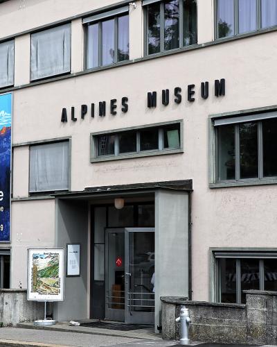 The image shows the exterior of a modern, multi-story building with a large banner advertising an exhibition titled "Schöne Berge" and prominent black lettering on the facade reading "ALPINES MUSEUM".