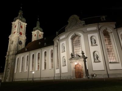A night photograph of a large, illuminated Baroque church featuring two prominent towers with green copper domes and a curved facade with statues in niches.