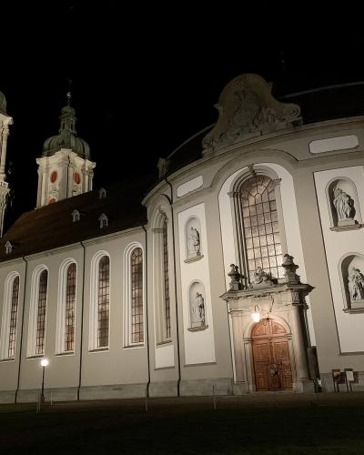 A night photograph of a large, illuminated Baroque church featuring two prominent towers with green copper domes and a curved facade with statues in niches.