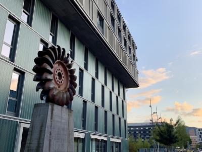 A low-angle shot of a modern multi-story building with green corrugated metal cladding and a large, rusted industrial turbine blade sculpture mounted on a concrete pedestal in the foreground, with a paved plaza and distant buildings under a twilig