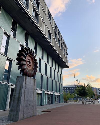 A low-angle shot of a modern multi-story building with green corrugated metal cladding and a large, rusted industrial turbine blade sculpture mounted on a concrete pedestal in the foreground, with a paved plaza and distant buildings under a twilig