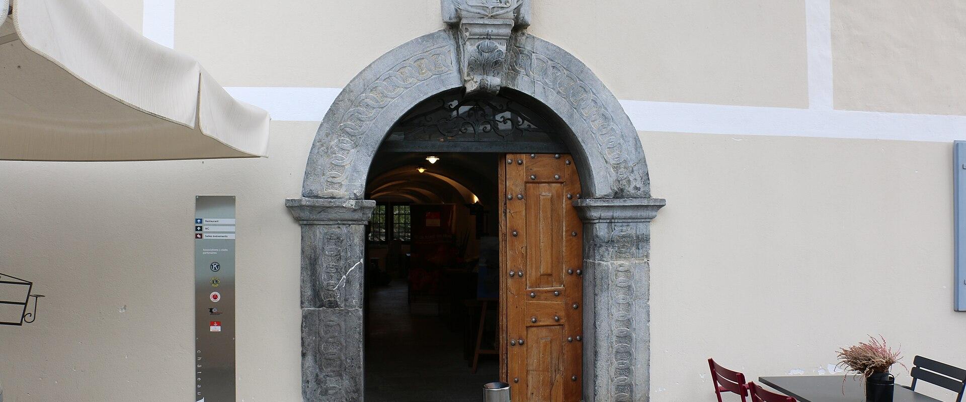 A close-up view of a stone arched entryway with a wooden door, set into a cream-colored building facade, featuring a vertical metal sign to the left of the arch.