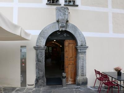 A close-up view of a stone arched entryway with a wooden door, set into a cream-colored building facade, featuring a vertical metal sign to the left of the arch.