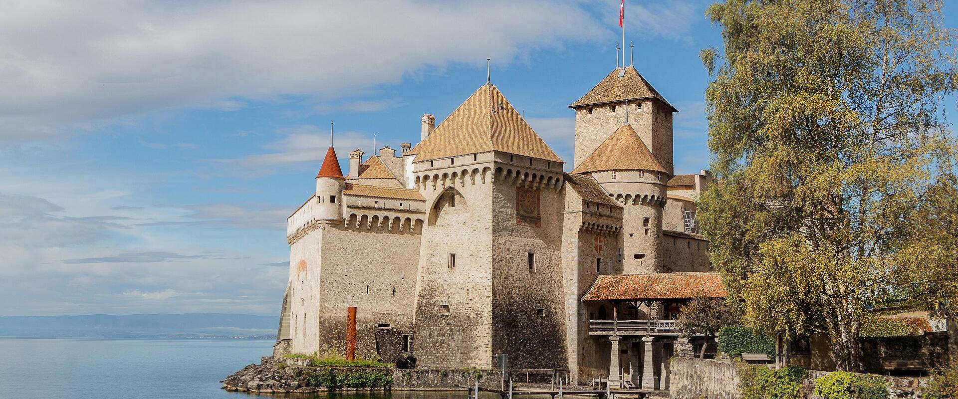 A close-up view of a medieval stone tower with a conical tiled roof and crenellations, attached to a high curtain wall, situated directly on the edge of a body of water with a distant shoreline visible.