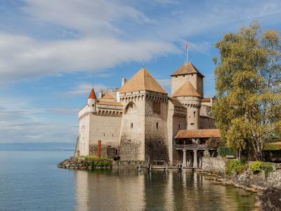 A close-up view of a medieval stone tower with a conical tiled roof and crenellations, attached to a high curtain wall, situated directly on the edge of a body of water with a distant shoreline visible.