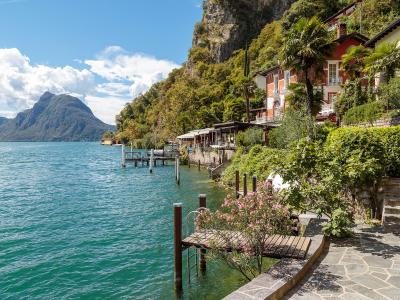 A lakeside promenade with stone paving, wooden docks, and terraced buildings with Mediterranean vegetation, overlooking a large lake with a distinctive mountain in the background.