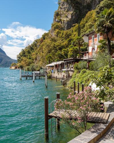 A lakeside promenade with stone paving, wooden docks, and terraced buildings with Mediterranean vegetation, overlooking a large lake with a distinctive mountain in the background.