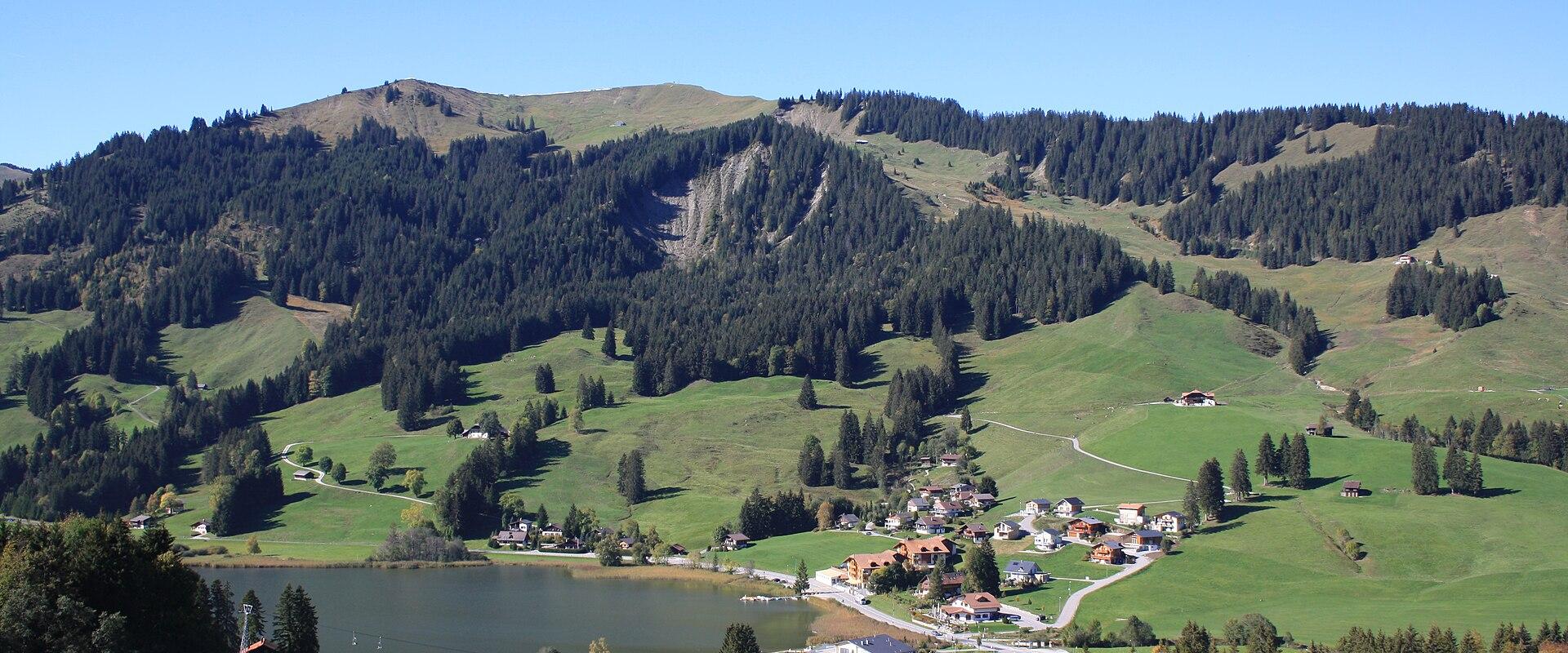 A wide landscape shot of a small lake nestled in a green valley surrounded by forested hills and a small village, taken during the summer or early autumn.
