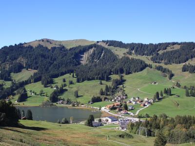 A wide landscape shot of a small lake nestled in a green valley surrounded by forested hills and a small village, taken during the summer or early autumn.