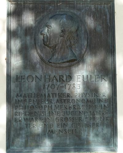 A close-up photograph of a rectangular bronze commemorative plaque mounted on a light-colored wall, featuring a relief portrait of Leonhard Euler in profile and inscribed text identifying him and stating he spent his youth in Riehen.