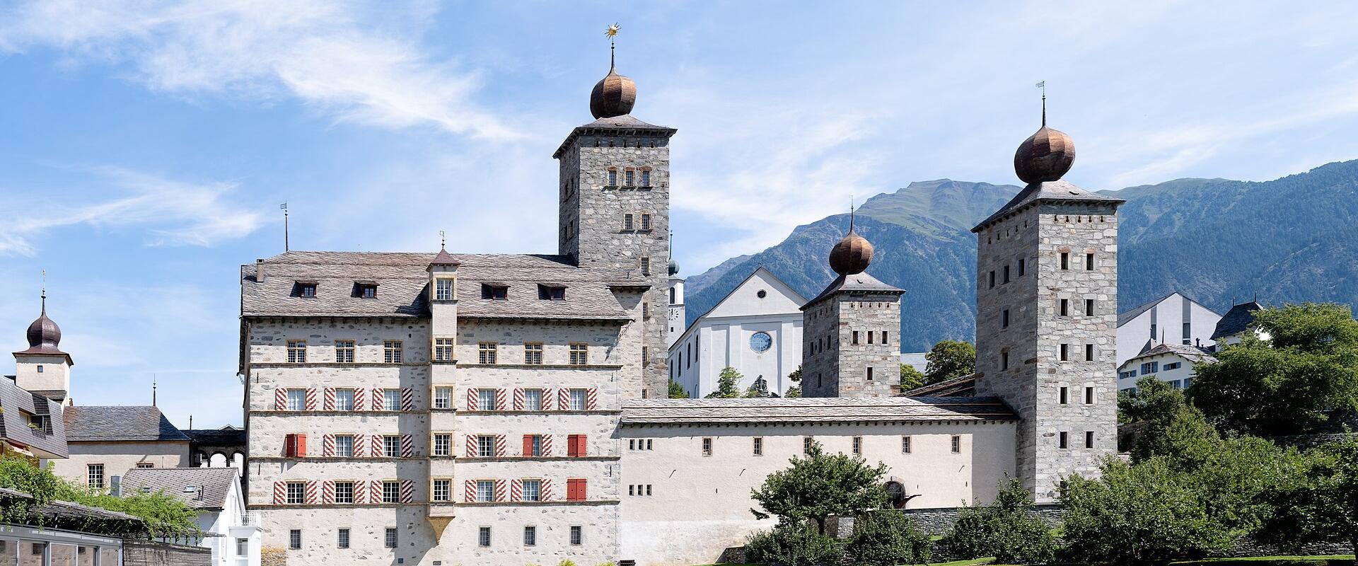 A large, fortified baroque palace complex with distinctive onion-domed towers, red and white striped shutters, and formal gardens, set against a backdrop of mountains.