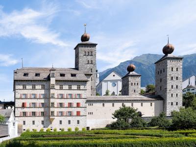 A large, fortified baroque palace complex with distinctive onion-domed towers, red and white striped shutters, and formal gardens, set against a backdrop of mountains.