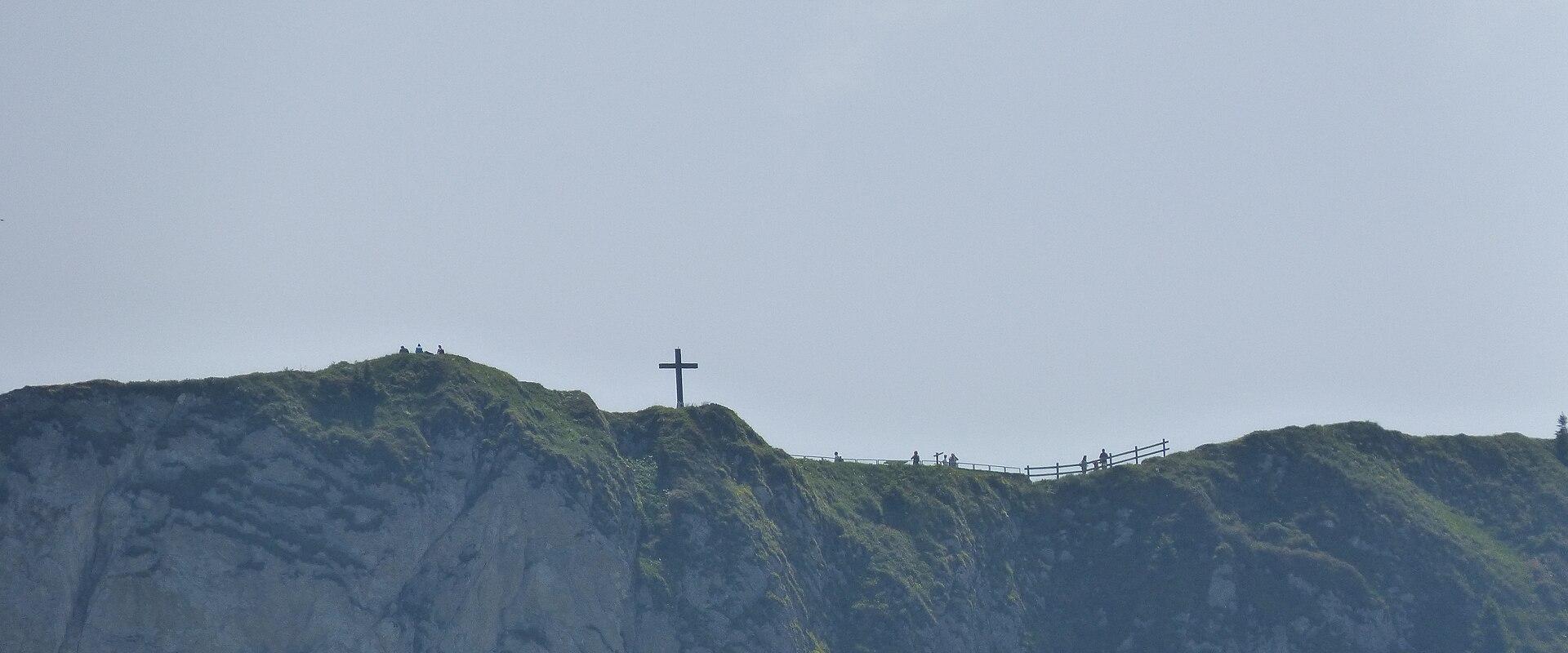 A rocky mountain ridge with a large cross on the summit, a wooden fence, and several people visible along the ridge against a pale sky.