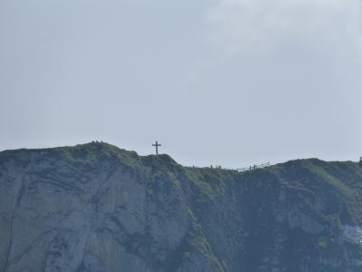 A rocky mountain ridge with a large cross on the summit, a wooden fence, and several people visible along the ridge against a pale sky.