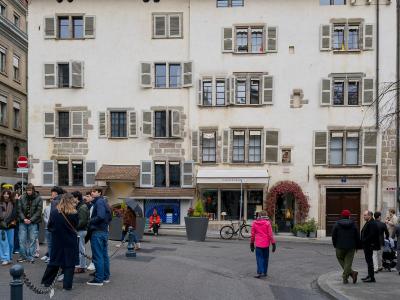 A street-level view of a large, cream-colored historic building with shuttered windows and a shop named "CAMILLE ARVEN" on the ground floor, with pedestrians gathered on the paved area in front.