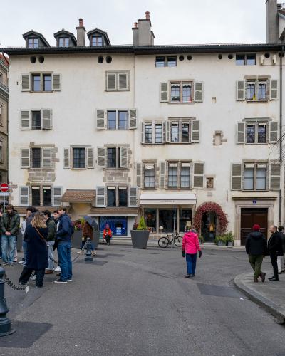 A street-level view of a large, cream-colored historic building with shuttered windows and a shop named "CAMILLE ARVEN" on the ground floor, with pedestrians gathered on the paved area in front.